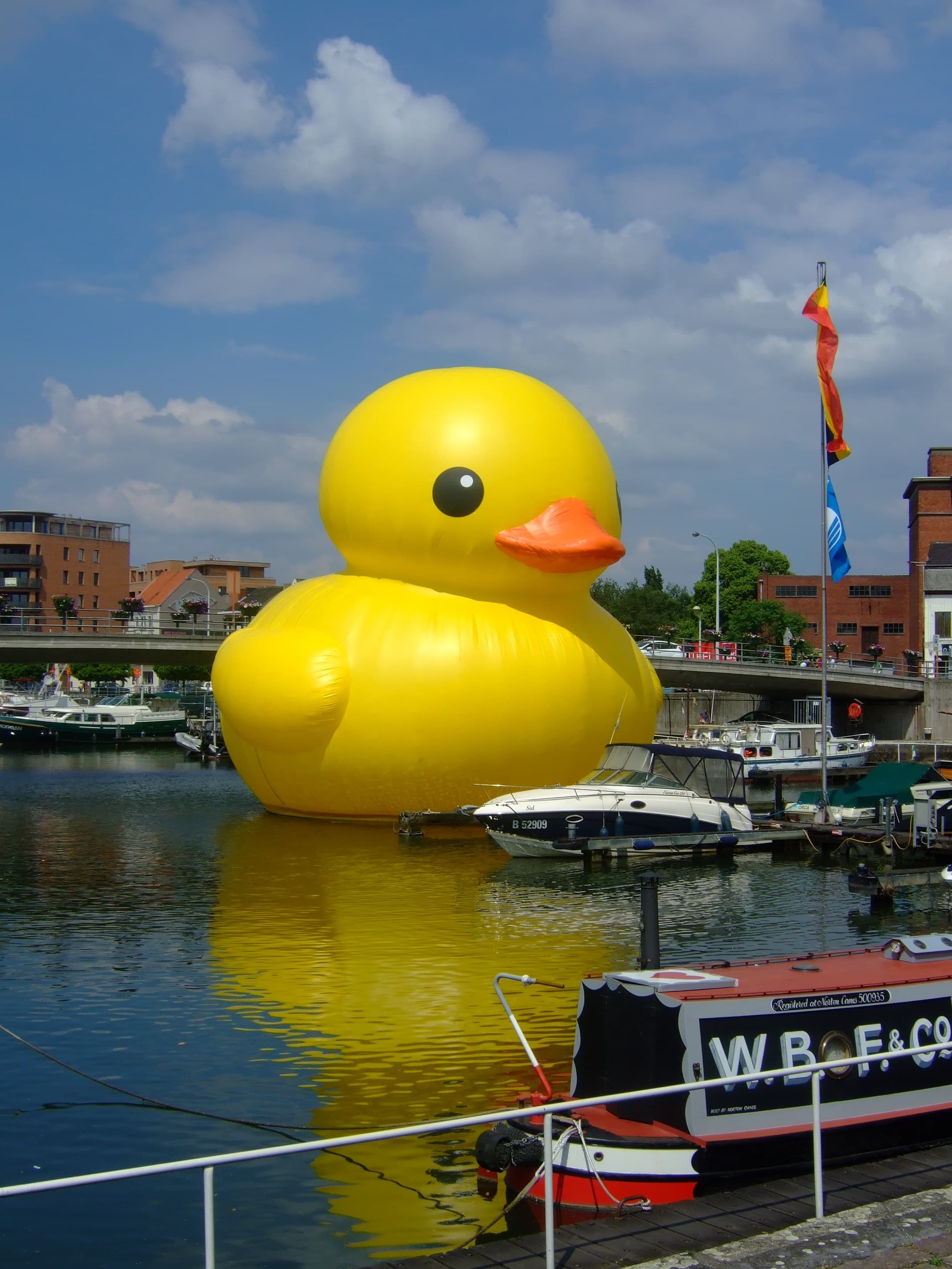 A giant inflatable rubber duck floating in a marina, moored boats and a bridge behind it